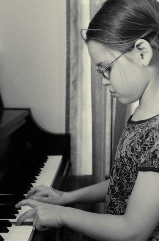 A young girl wearing glasses concentrates on the keys as she plays an upright piano.
